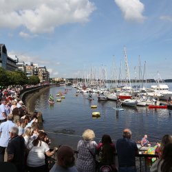 Crowd gathered along a waterfront watching boats and kayakers in a busy marina under a sunny sky.