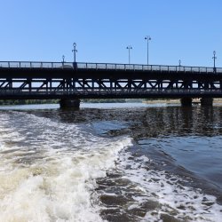 Boat wake on the River Foyle passing beneath the Craigavon Bridge against a clear blue sky.