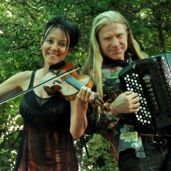 Two musicians stand outdoors in a leafy, sunlit forest. A smiling woman in a sleeveless black dress plays a violin in the foreground, while beside her a man with long blond hair plays a button accordi