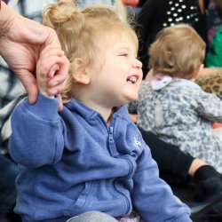 A young baby sat up holding their carers hand laughing.