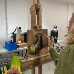 Female painter standing at easel paints still life of jug and a squash