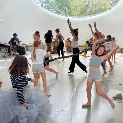A picture of people dancing and playing with fabrics. There is someone in the background playing music and a child in the foreground wearing many net skirts