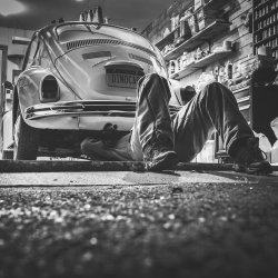 A mechanic lies on their back, looking underneath an older car