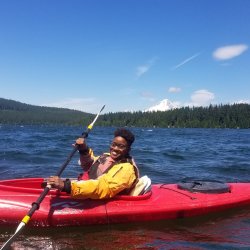 A person wearing a P-F-D (lifejacket) paddles a kayak on a calm lake with Mt. Hood in the background