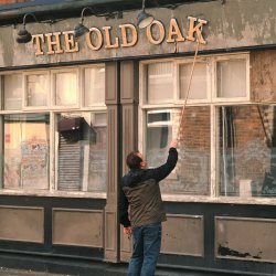 A still from the film featuring an old pub front, with a sign that reads 'THE OLD OAK'. The final letter K is falling, and a person is trying to fix it with a long stick