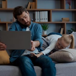 Dad on his phone and laptop trying to work while his daughter tries to play with him and his computer.
