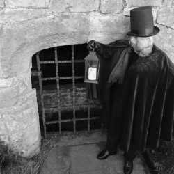 A performer from Time Will Tell Theatre Company. stands with a lantern outside a metal grate in a stone wall