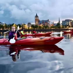 Kayakers on a river at dusk with city lights.