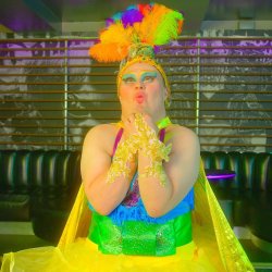 Performer in colourful drag costume poses in a nightclub, wearing a sparkling headdress with bright rainbow feathers, dramatic blue eye make-up, gold gloves and a vivid green and blue outfit.