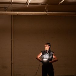 A performer stands against a large brick wall in a dim, warm-toned industrial space. Wearing a dark sleeveless outfit, they hold a glowing circular light at their waist, illuminating their face and to