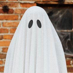 Spooky ghost standing in front of an abandoned building - the ghost is a person wearing a white sheet with black cardboard ovals stuck to the front for eyes