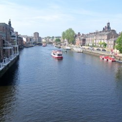 View along the River Ouse in York on a bright day, with a sightseeing boat on the water, historic riverside buildings, trees and boats moored along the bank. The photograph is taken from Skeldergate B