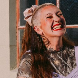 Colour photograph of folk singer Jennifer Reid. Her hair is shaved at the sides with a long, red ponytail at the back. Jennifer is wearing red lipstick and laughing.