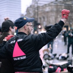 A group of people protest outdoors in a city; one person raises a red gloved fist, wearing a jacket with a pink triangle and the words "ACT-UP LONDON STILL NOT SILENT"