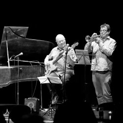 Black-and-white photo of a jazz trio performing on stage. A pianist, bassist and trumpeter play beneath spotlights, with music stands, microphones and audience silhouettes in the foreground.
