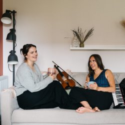 Rachel Campbell and Holli Scott are sitting with their feet up facing each other on a sofa. There is a fiddle next to Rachel and an accordion next to Holli. They are both holding a mug and laughing.