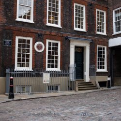 Exterior view of Dr Johnson’s House in Gough Square, London—a Georgian brick townhouse with white-framed sash windows and a dark green front door, accessed by a short flight of steps. A round commemor