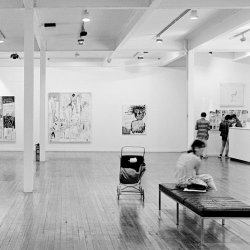 A black and white photo of an exhibition space and a child's buggy in the Frutimarket in 1984. There is a buggy in the space and people milling around the Basquait exhibition