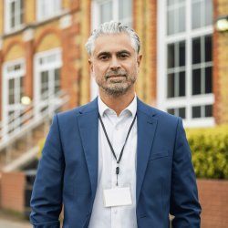 Man in suit wearing a lanyard, standing outside a school building