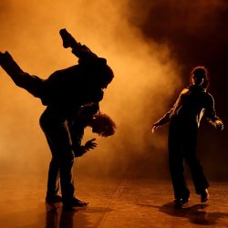Three contemporary dancers in an improvised moment: one lifting another in motion while a female dancer moves behind them, all set against warm orange light and atmospheric haze