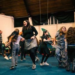 A lively group of adults and children enjoying a ceilidh dance in a bright hall, full of energy and joy.