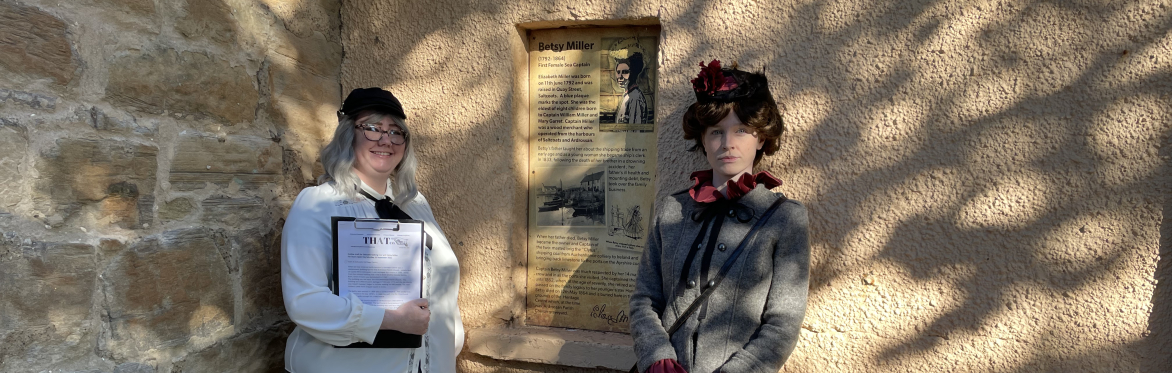 Two women, one dressed as Betsy Miller standing at the Captain Betsy Miller information board.