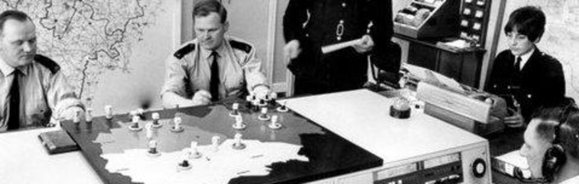 Black and white image of a police control room. There are maps on the walls and three policeman and one police woman sit around the control table which has various gadgets. Another officer is standing
