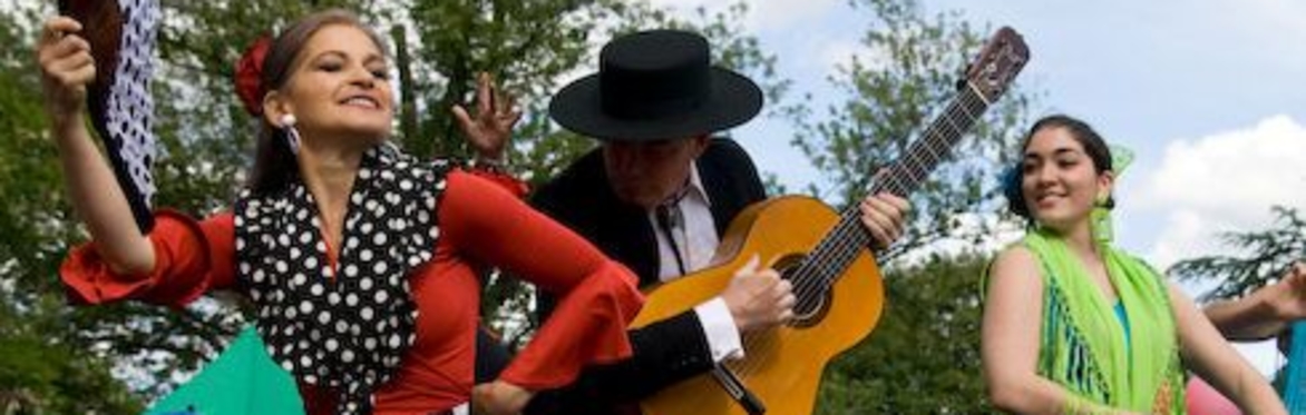 Performance action shot, set on an open-air stage: two flamenco dancers in brightly-coloured spotty dresses are accompanied by a flamenco guitarist in a dark suit and a Spanish wide-brimmed hat.