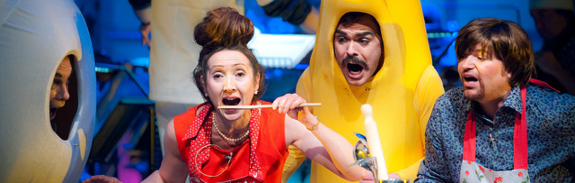 A person dressed as an egg, a women eating with a spoon and man dressed as a banana and someone in an apron, sit alongside each other