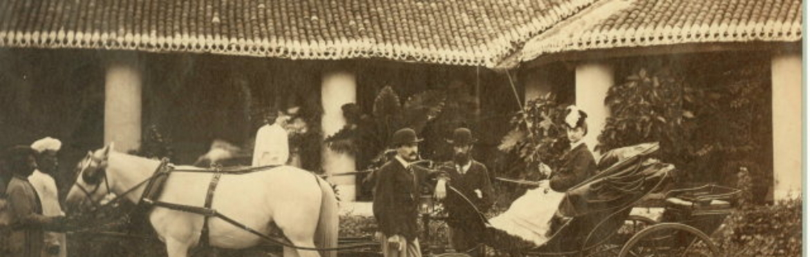 A British Colonial family outside their house in India. A white woman is sitting in an open carriage attached to a white house. There are two Indian servants by the horse's head. Many in a bowler hat