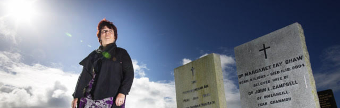 Fiona dressed in dark jacket and legging with a flowery purple dress, stood next to the white gravestone of Margaret Fay Shaw with a blue sky in the background and green grass in the foreground
