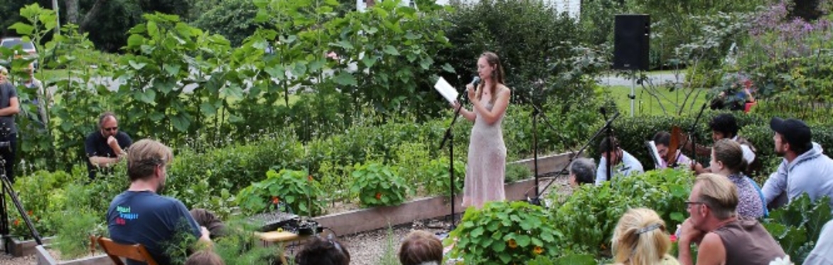 Audience seated in a garden listens to a performer reading from a microphone, with lush green plants in the foreground and forested mountains and a white house in the background. An intimate outdoor p