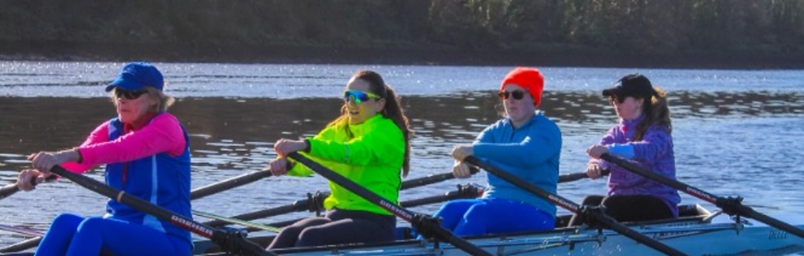 Four people in vibrant athletic gear row a boat across a calm, sunlit river.