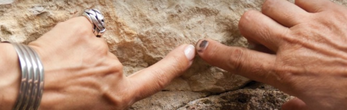 Two hands, one white toned and one brown toned have index fingers outstretched, touching. Both hands seem to be feeling together a craggy sandy coloured rockface.