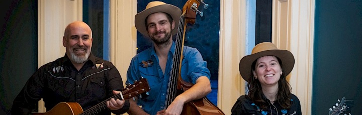 The three members of Good Guy Hank standing in front of a window with hats on holding their instruments.