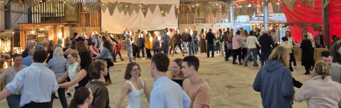 Dancers around the barn circling in groups of four. Bunting hangs from the barn roof and a cheerful red sail is along one wall. The bar and the food counter are lit up at the other end of the barn.