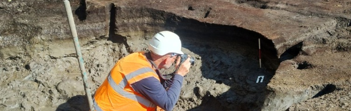 An archaeologist in an orange safety vest photographs a muddy excavation trench, documenting exposed soil layers and a dark cavity in bright sunlight.