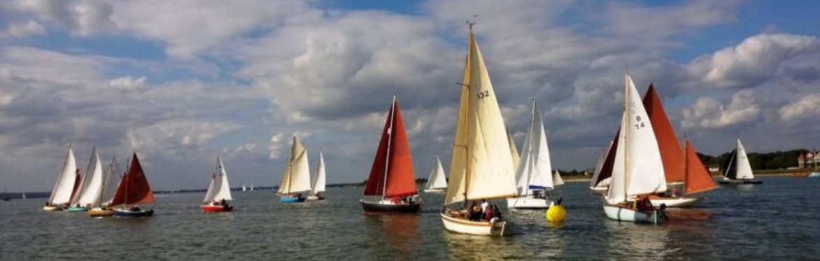A fleet of traditional sailing boats with white, cream and red sails race across calm estuary waters beneath a dramatic sky filled with large clouds.