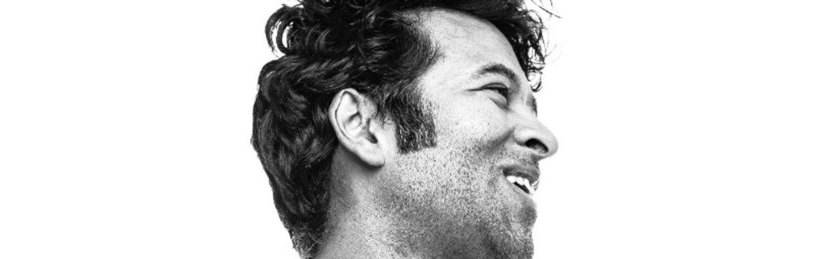 Black-and-white portrait of composer and performer Paul Pinto. He stands in profile, facing to the right, with his arms casually crossed and a slight smile on his face. He wears a plain white T-shirt