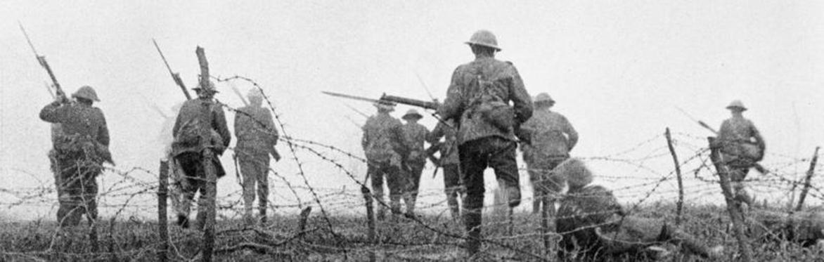 Black-and-white photograph of First World War soldiers carrying rifles as they advance through barbed wire across a battlefield.