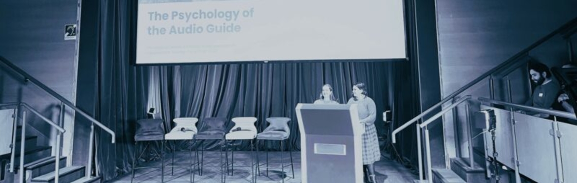 Two women are standing on a stage behind a lectern, there's empty chairs on the stage and a big screen behind them