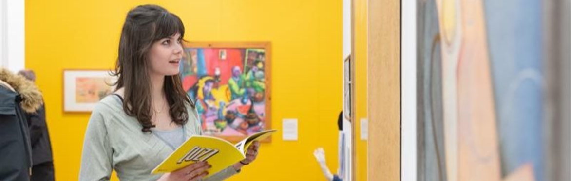 A person holding a yellow catalogue in front of a yellow wall with paintings.