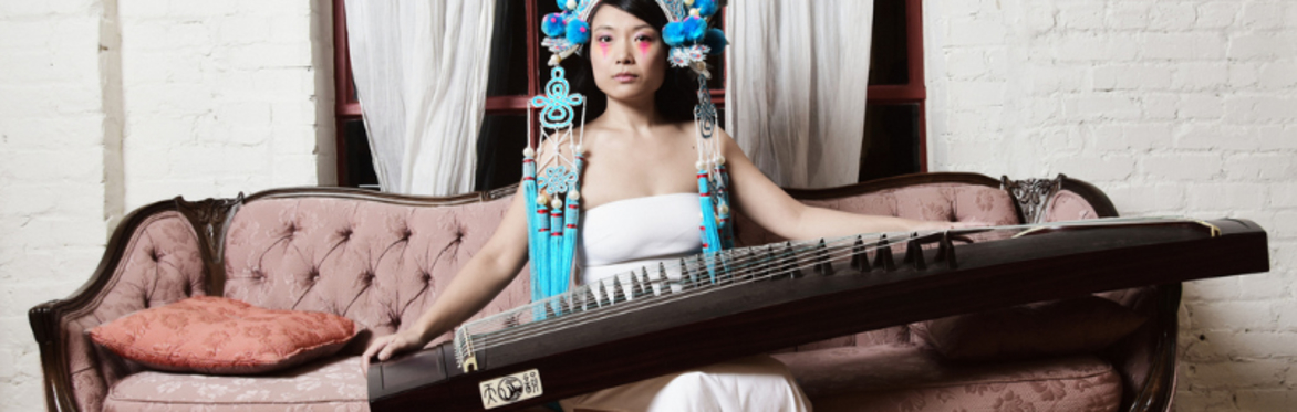 Wu Fei in a traditional Chinese opera headdress and white dress plays the guzheng while seated on a pink sofa.