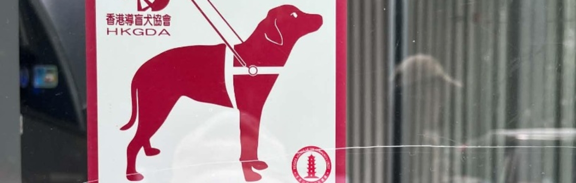 Red and white sticker from Hong Kong Guide Dog Association on a glass door that shows a drawing of a guide dog with the caption "Guide dogs welcome". Blue and white wheelchair symbol sticker is below.