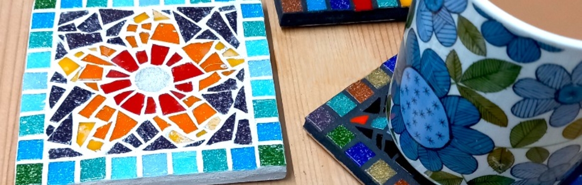 Several handmade coasters, one has a mug of tea on it. The main coaster featured has an orange mosaic tile flower and a blue mosaic tile border,