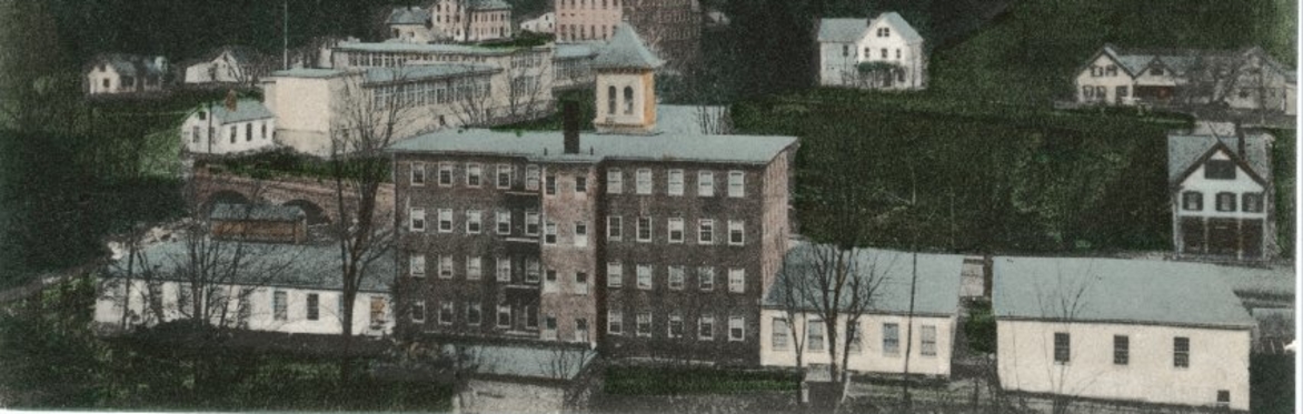 Colorized vintage postcard showing a complex of brick and clapboard mill buildings in a green, hilly landscape.
