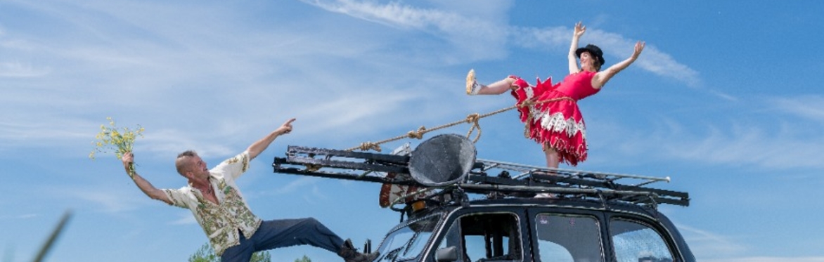 2 circus peformers balance on top of a black cab, one holding a bunch of flowers