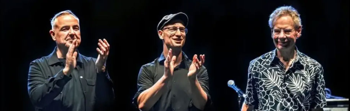 **Alt text (under 200 characters):** Three musicians stand on stage acknowledging applause. Two clap while the third smiles, with drums and stage equipment behind them under concert lighting.