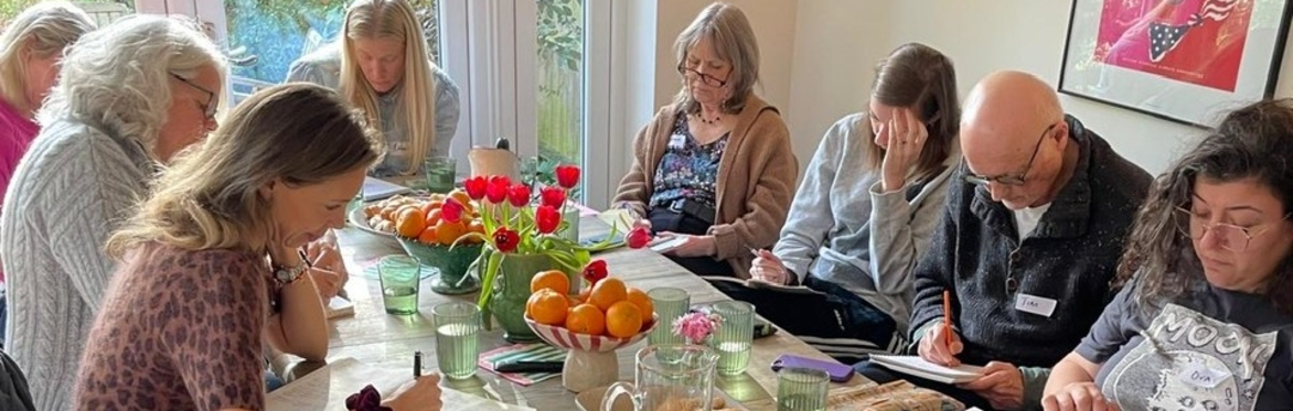 A group of writers sit around a table with flowers and fruit, deep in concentration as they write