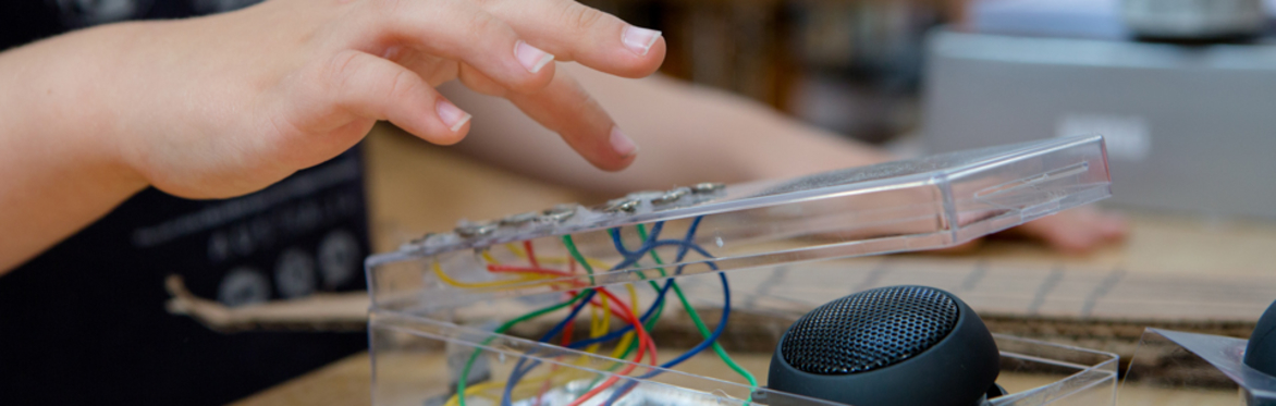 Close-up of a hand interacting with a DIY electronics project in a clear plastic case containing coloured wires and a small speaker. The image represents hands-on digital making, technology learning,